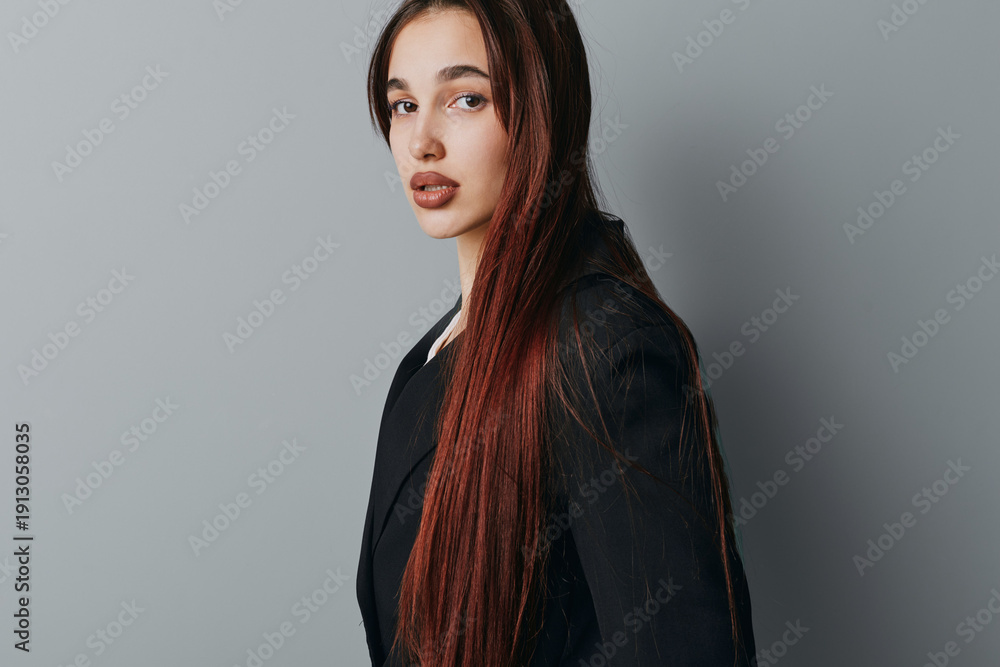 Naklejka premium Young woman with long straight hair wearing black blazer posing and looking at camera with neutral expression on gray background in studio portrait. Female model with natural makeup.