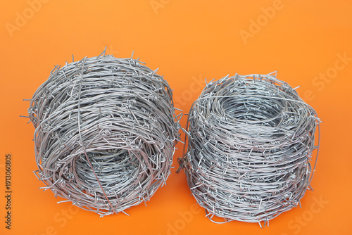 Two rolls of galvanized barbed wire placed side by side on a bright orange background. Industrial metal coils with sharp spikes symbolize security, protection, restriction, and construction materials 