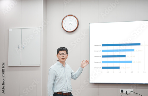 Fotografija A bespectacled man standing in a classroom pointing toward a blackboard while ex
