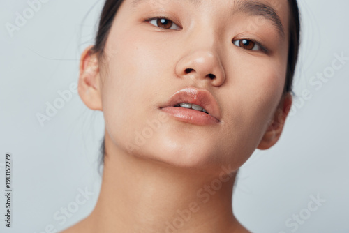 Portrait of a young woman with clear skin and natural makeup looking at camera with neutral expression. Closeup face with smooth complexion and glossy lips on neutral background.