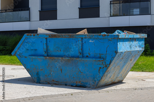 Large blue metal construction dumpster placed near modern residential building on sunny day, industrial waste container for renovation debris and urban cleanup concept