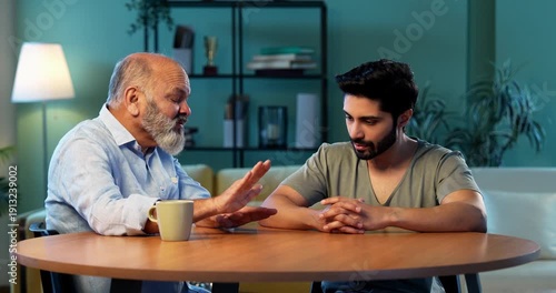 Indian senior father advising young son during serious life discussion while sitting at table in modern living room at home, experienced parent guiding student child about responsibility, future and v