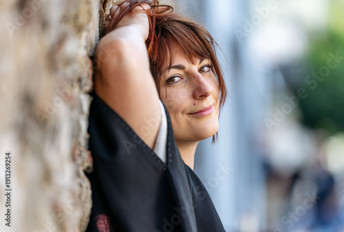 Canvas Print Young woman with auburn hair leans against a textured stone wall, smiling softly