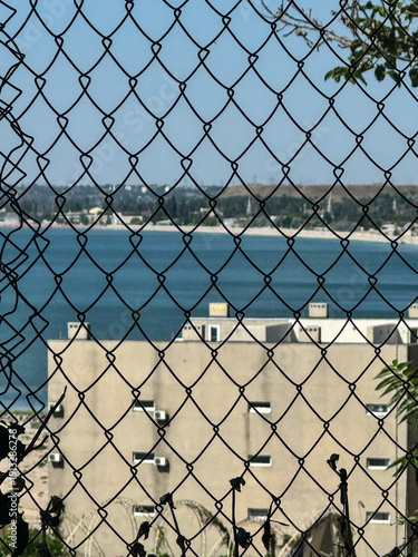 View of Sea and Building Through Chain Link Fence, showcasing urban landscape and coastal beauty, creating a sense of enclosure and observation