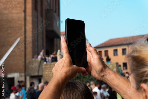 Photos Person taking photos with smartphone at a public event in a historic city, crowd in background, mobile technology and urban outdoor lifestyle