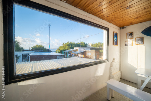 A large black-framed window offers a bright view of a coastal village under a blue sky. The interior features white walls, rustic wooden ceilings, and a clean, minimalist aesthetic.