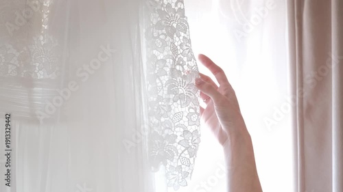 Close-up of a white bridal gown featuring intricate floral lace and sheer tulle fabric hanging near a window.