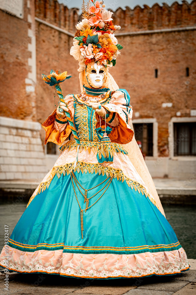 Obraz premium Venice, Italy - February 09, 2026: Elegant masked figure in vibrant blue and orange costume adorned with flowers at a historical venue during a festive celebration