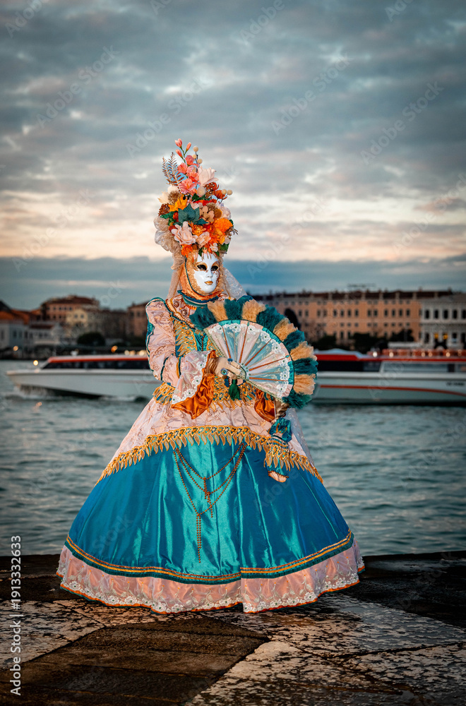 Naklejka premium Venice, Italy - February 09, 2026: Colorful masked performer in elaborate costume holding a fan by the water during a vibrant carnival celebration at sunset