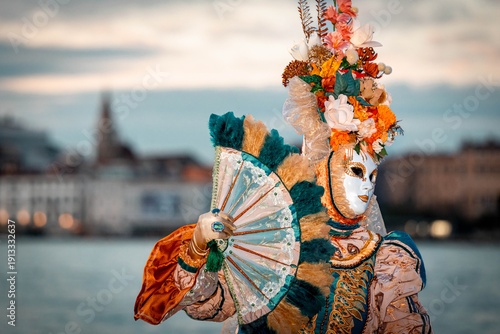 Venice, Italy - February 09, 2026: Colorful masked figure in elaborate costume holding a fan during a carnival celebration with historic buildings in the background