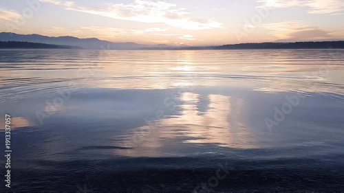 A serene water surface with expanding ripples reflecting a colorful sky and distant hazy mountains at dawn The suns glow illuminates the horizon