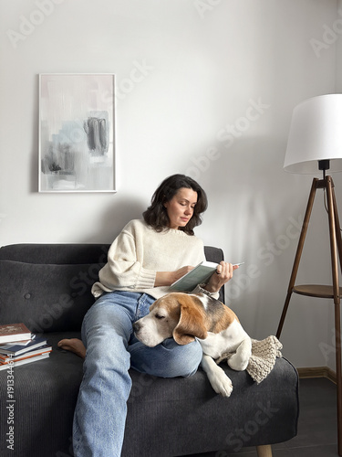 Photography Cozy home education young woman reading a book on a sofa with her beagle dog at home