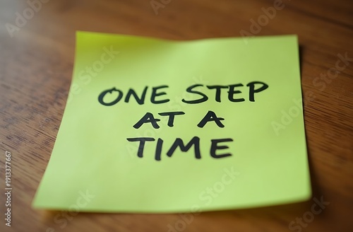 green sticky note placed on a wooden desk with the phrase ‘ONE STEP AT A TIME’ written in neat black handwriting