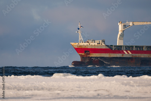 Cargo ship sailing through the frozen Baltic Sea with icy foreground, industrial vessel navigating cold winter waters under cloudy sky