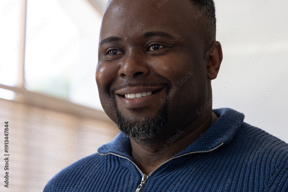 Obraz premium African American man smiling wearing blue ribbed zip sweater facing window blinds at home