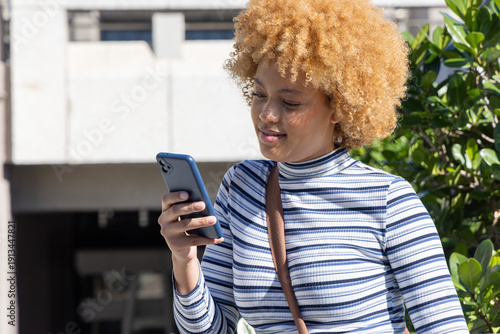 African American woman using blue phone outdoors near building in striped top with brown bag strap