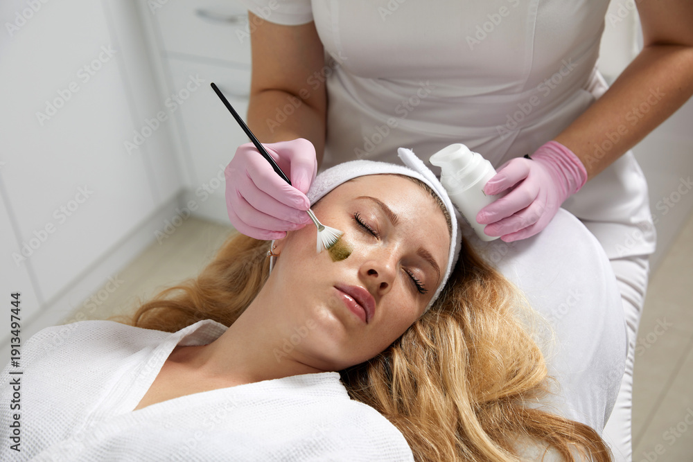 Naklejka premium Cosmetologist applying a mask from clay to the face of a beautiful woman. Spa treatment and face care in the beauty salon