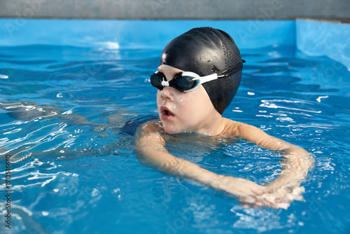 Preschool boy learning how to swim in a pool wearing swimming cap and goggles