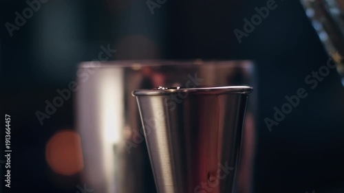 Close-up of dark alcohol being poured into a metal jigger in a dimly lit cocktail bar.