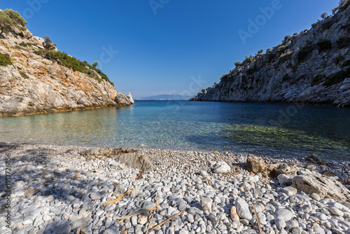 View of a small, idyllic and empty pebble stone beach in a cove surrounded by rugged and hilly coastline by the Aegean Sea on Kalymnos island in Greece on a sunny day.