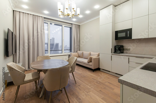 Naklejka Bright living room with compact kitchen and panoramic city view. Beige furniture, gold accents and wooden flooring create a cozy yet modern ambiance.