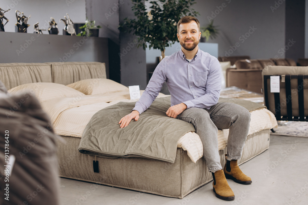 Obraz premium Happy man in a furniture showroom sitting on a comfortable bed, touching the bedding while shopping for home decor