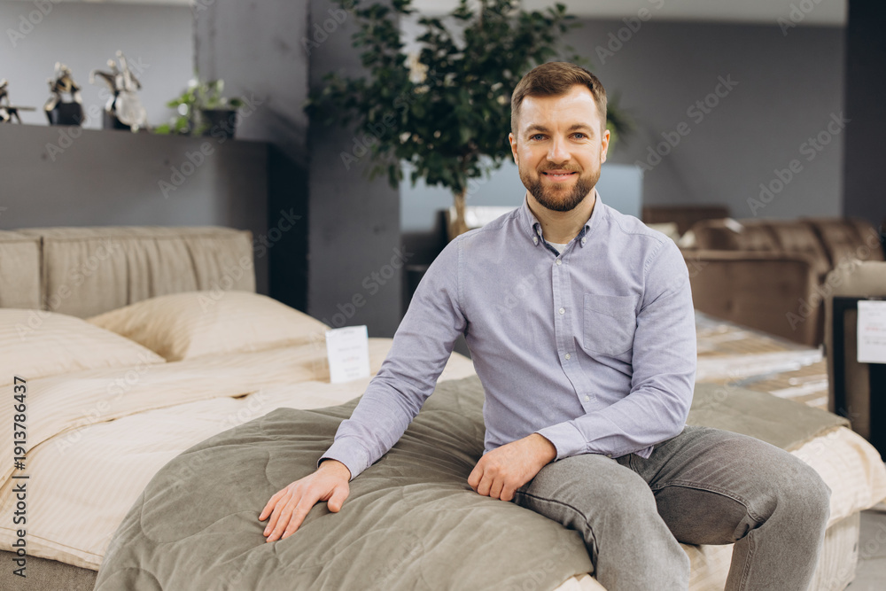 Fototapeta premium Smiling man sitting on a bed in a furniture store showroom, trying out mattresses, shopping for home interior items