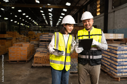 Two warehouse workers wearing safety gear discuss logistics while holding tablets, surrounded by stacks of boxes in the storage area.