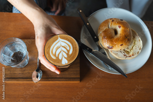 Top view of hand 's holding hot latte coffee with latte art in a glass and bread on wooden table, flat lay concept.