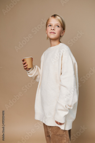 Photos Authentic lifestyle portrait of a young girl in a cozy oversized sweater, holding a cup, conveying playful schooldays and simple everyday moments in a calm studio setting
