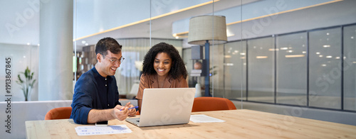 Smiling team professional employees working together on computer discussing online technology data solutions at work. Two young busy diverse coworkers business people using laptop at office meeting.