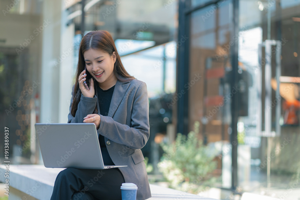 Fototapeta premium Businesswoman talks on phone, updates tasks, and plans milestones on a laptop. Online business, technology, in front of a modern office building.