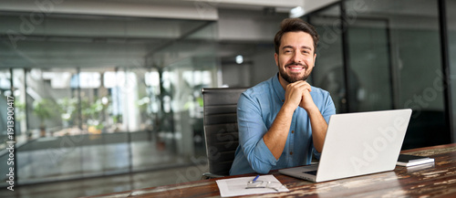 Happy professional business man company employee, young entrepreneur, smiling latin businessman working on laptop computer technology looking at camera working in office sitting at desk, portrait.