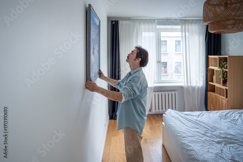 Obraz na płótnie Interested man hanging framed picture on white wall in bedroom