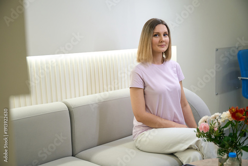 Smiling young pretty woman waiting in hall of clinic, sitting on sofa