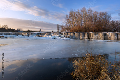 Winter weir at Cvrcovice with ice cascades in warm light. Long exposure creates a silky water effect amidst a frozen natural landscape.