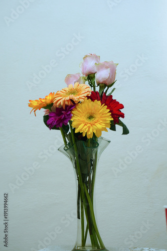 A close-up still life featuring three vibrant gerbera daisies in shades of orange and yellow, elegantly arranged in a modern wavy glass vase against a neutral light background