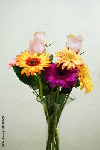 A close-up still life featuring three vibrant gerbera daisies in shades of orange and yellow, elegantly arranged in a modern wavy glass vase against a neutral light background