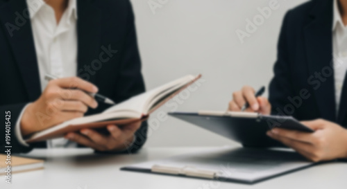 Soft focus blurred photo of Two business professionals in dark suits reviewing documents and a notebook during a meeting, with a blurred white background.