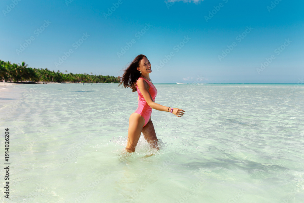Naklejka premium Joyful pretty woman in pink swimsuit running in blue ocean water, enjoying summer tropical vacation and sunny day