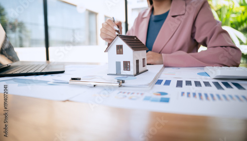 A businesswoman reviews financial charts and documents beside a small house model, analyzing real estate investment and mortgage planning at her desk.