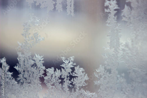 The photo captures delicate frost patterns on the window glass. The white ice crystals resemble tree branches against a blurred, light background.
