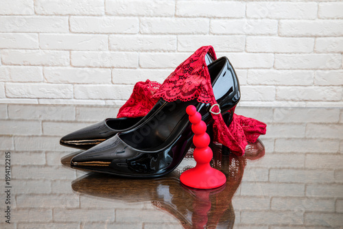 Black high heel shoes with red lace panties and red anal plug on glass table against white brick wall