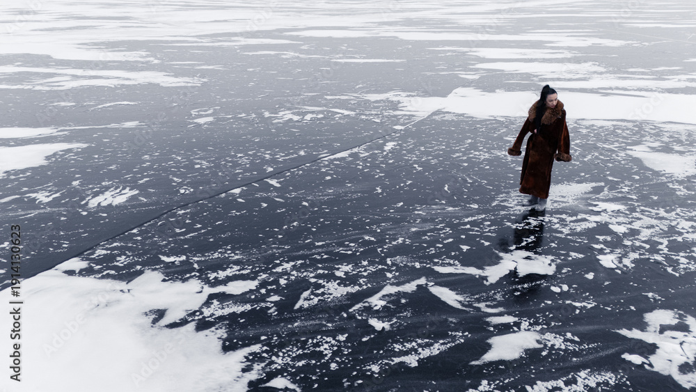 Fototapeta premium Woman in long fur coat walking on dark ice of vast frozen lake winter landscape