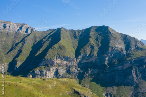 Steep grassy slopes and dramatic rocky cliffs rise under clear blue skies in the Pyrenees near Gavarnie Gedre. Strong sunlight casts deep shadows, highlighting the rugged textures and contours of the