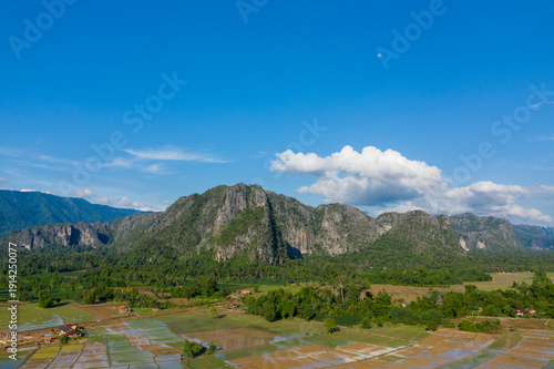 Expansive view of rugged limestone mountains rising above flooded rice fields and scattered rural houses, set beneath a vivid blue sky with scattered clouds. The landscape highlights the layered