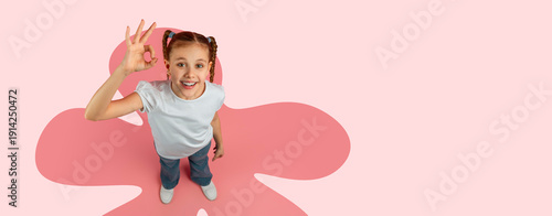 A young girl stands in a bright space with a large pink design on the floor. She smiles widely and shows an OK hand gesture. The scene captures her joy and playful spirit.