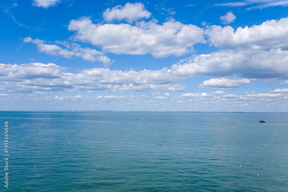 Fototapeta premium A wide view of the calm Atlantic Ocean stretches beneath a sky filled with soft, layered clouds. The tranquil scene features open water, subtle ripples, and a distant boat, evoking a sense of space