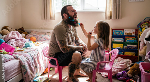 Laughing tattooed father sits on small pink chair while daughter puts hair clips in his beard in messy playroom