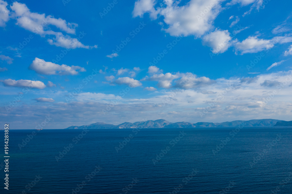 Obraz premium Vast blue ocean stretches toward a hazy mountain range on the horizon under a sky filled with scattered clouds. The tranquil scene captures natural gradients, open space, and a sense of calm.
